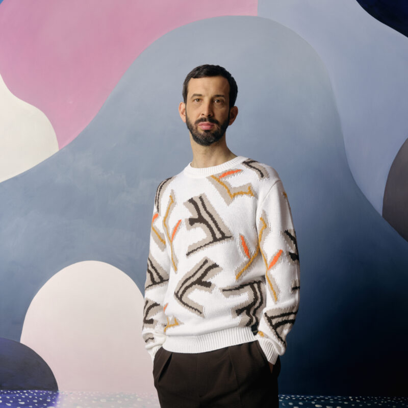 Man with dark hair and beard in a patterned white jumper and dark trousers, against a wall with pink, grey and white shapes on it.