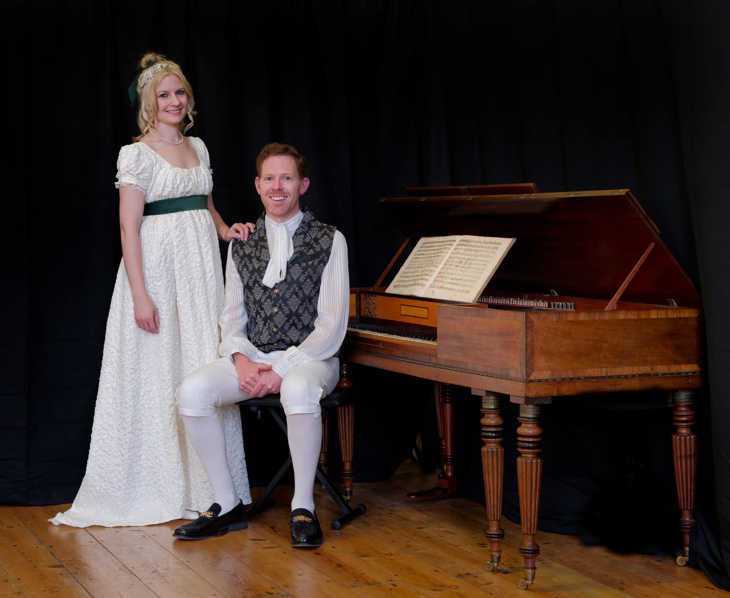 A woman standing in a white dress and a man sitting in a waistcoat by a piano
