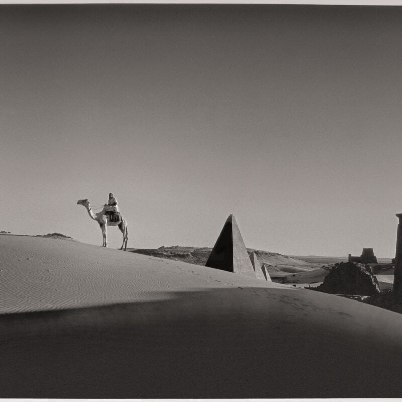 Black and white photograph of a desert with sand dunes and pyramids in the back ground. In the center of the image is a man riding a camel.