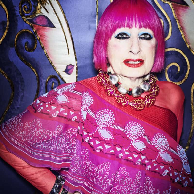 Photograph of Zandra Rhodes, a woman with a pink bob and vivid eye make up. Wearing pink patterned clothing, should against a blue patterned background.