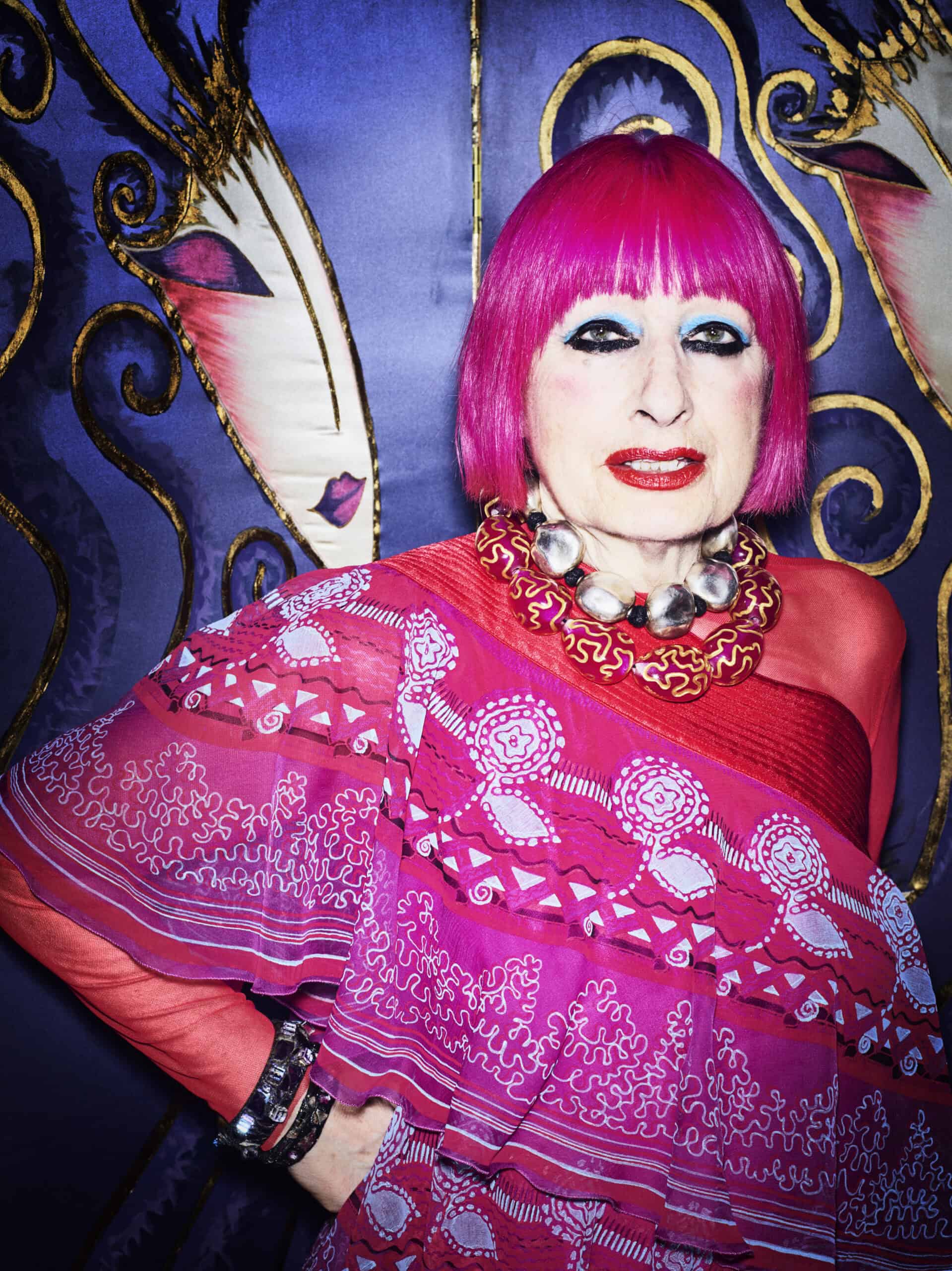 Photograph of Zandra Rhodes, a woman with a pink bob and vivid eye make up. Wearing pink patterned clothing, should against a blue patterned background.