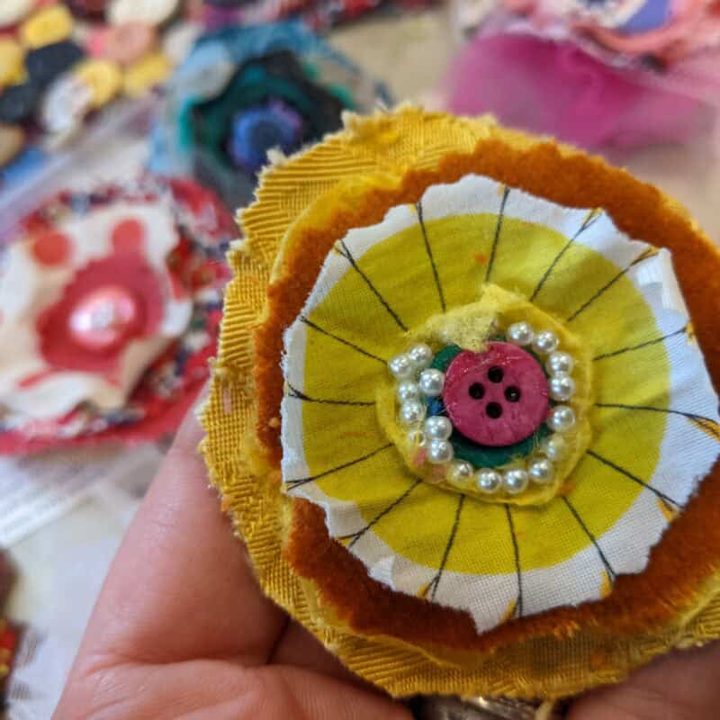 A fabric flower brooch with several layers of yellow, red and white felt, and a red button in the middle surrounded by pearls