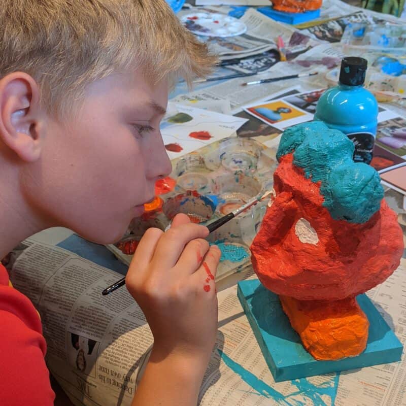 A blond haired boy carefully painting a blue and red model head with a paintbrush