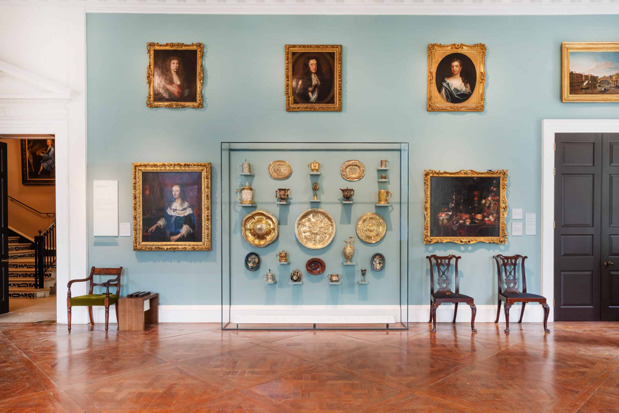 Photo showing a wall display in the museum with portrait paintings in gold frames, and ceramic objects inside a glass case