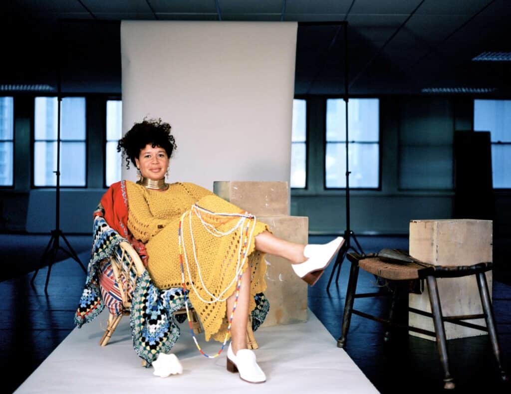 The artist Alberta Whittle sits in a chair, wearing a yellow dress