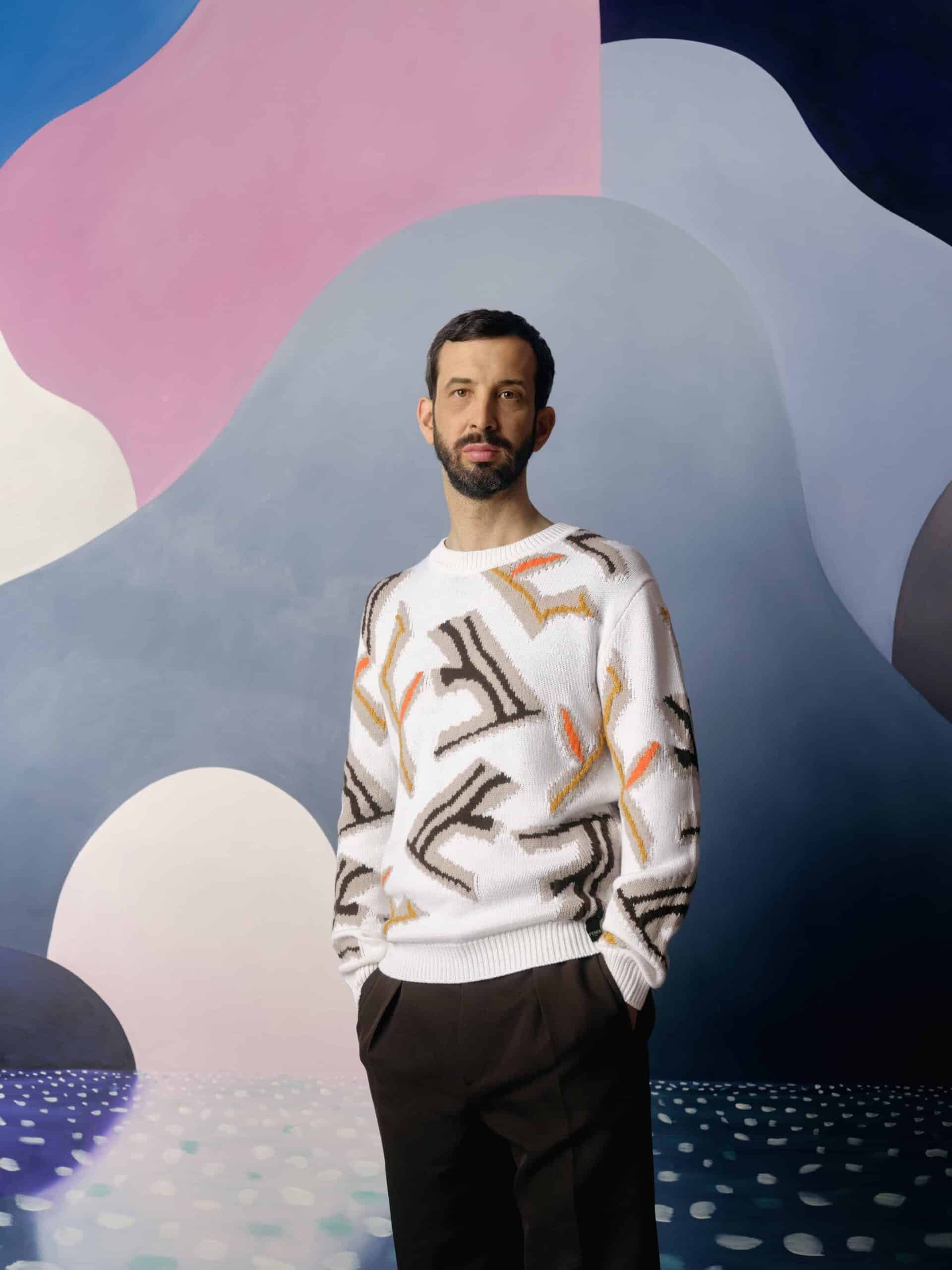 Man with dark hair and beard in a patterned white jumper and dark trousers, against a wall with pink, grey and white shapes on it.