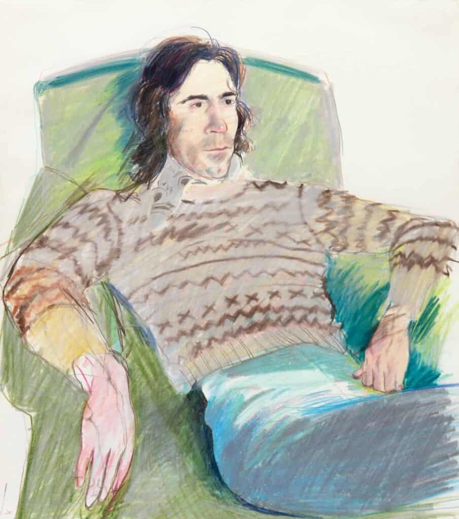 A man with long dark hair in a knitted sweater and jeans, relaxing in a green armchair