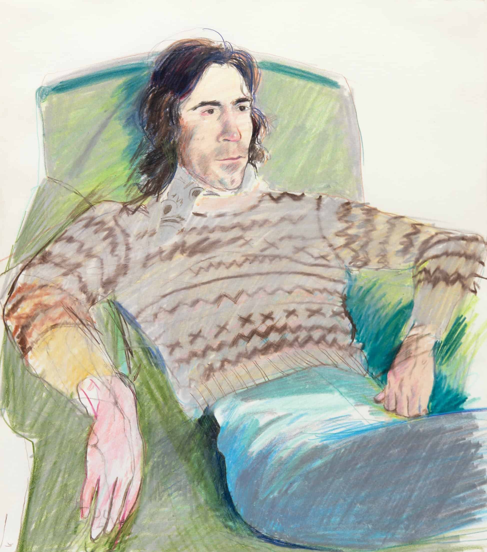 A man with long dark hair in a knitted sweater and jeans, relaxing in a green armchair