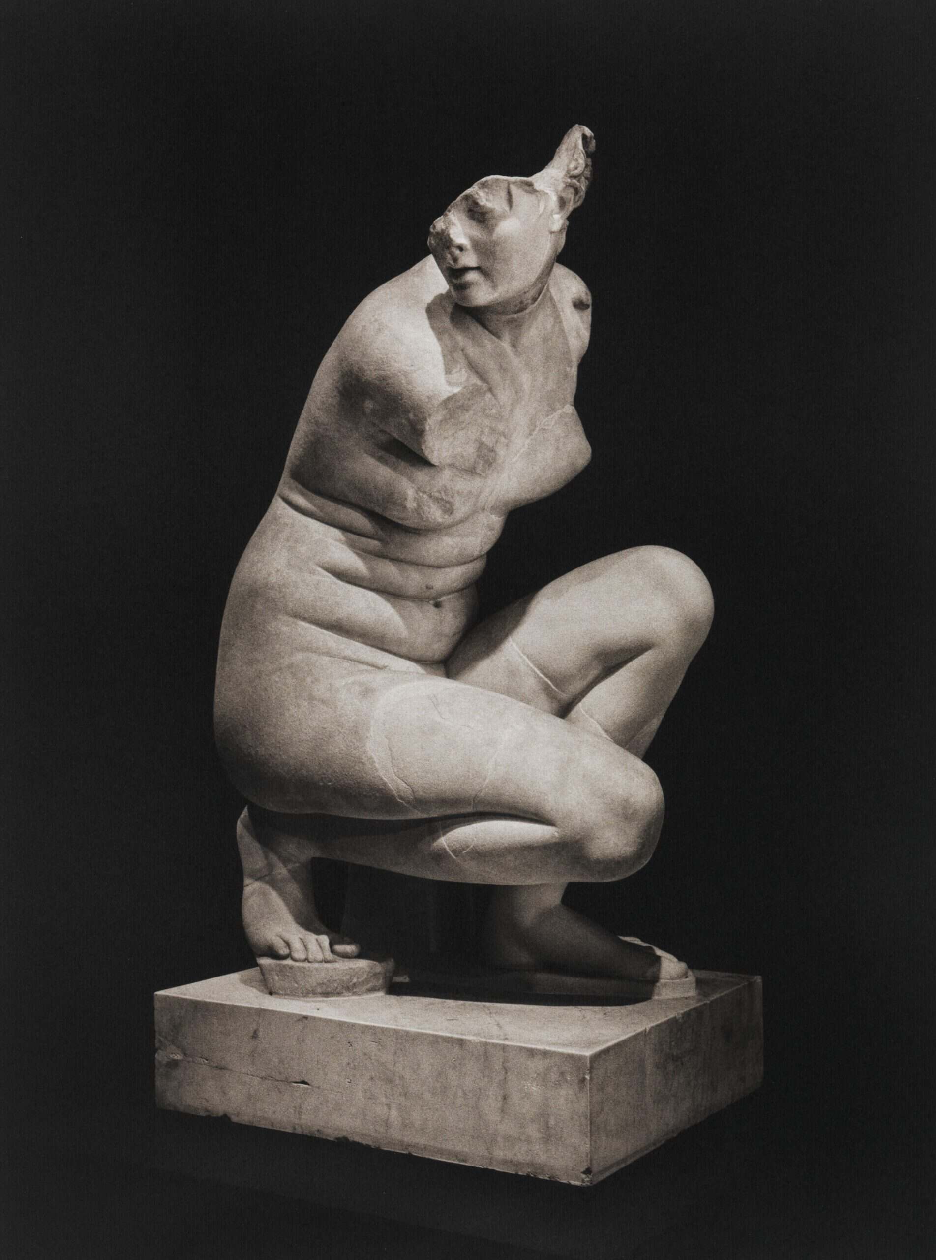 A sculpture of a female nude crouching with no arms and part of her head missing on a very dense black background