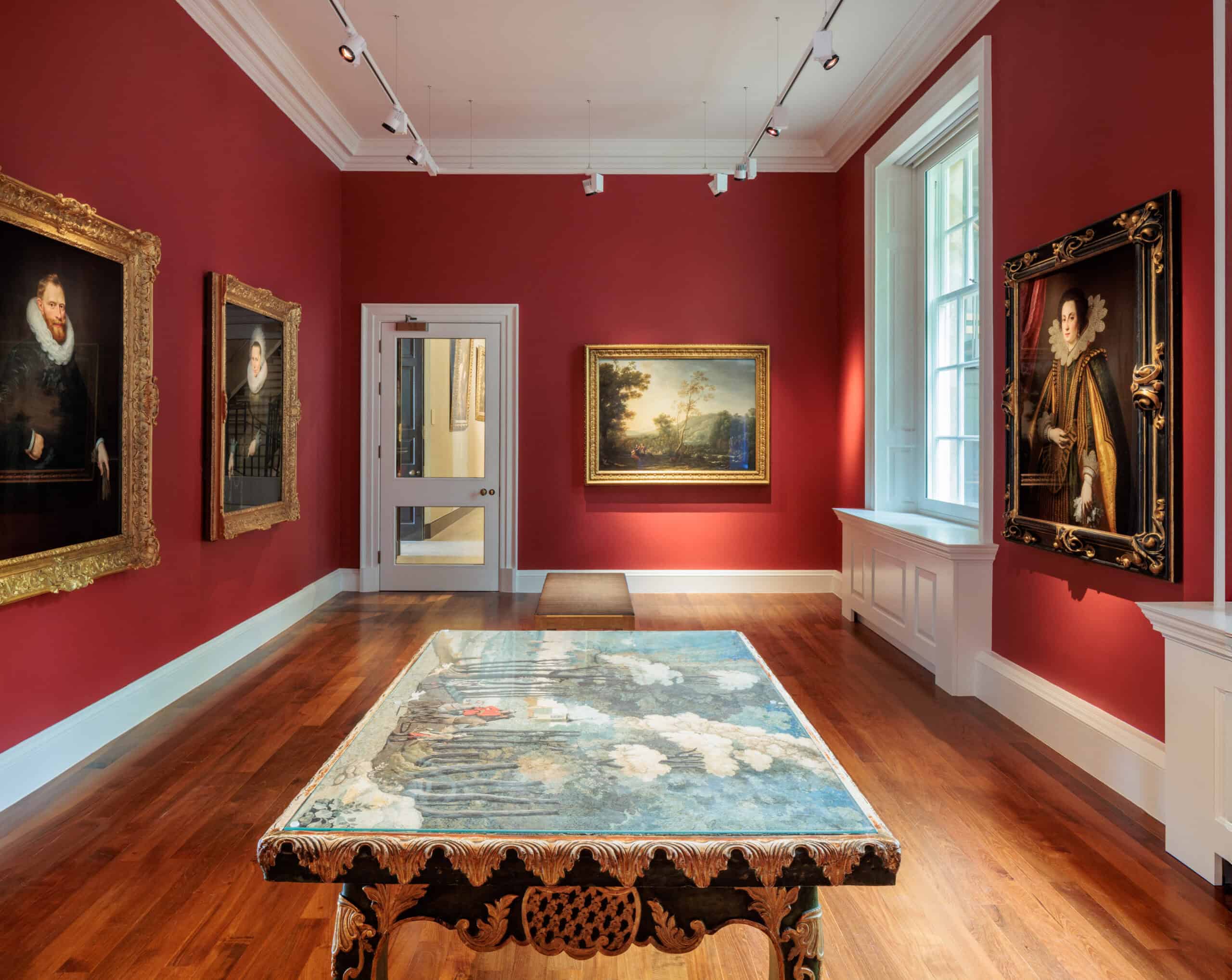 Photo showing a gallery with dark red walls, paintings in gold frames and a large table in the foreground with a decorative surface