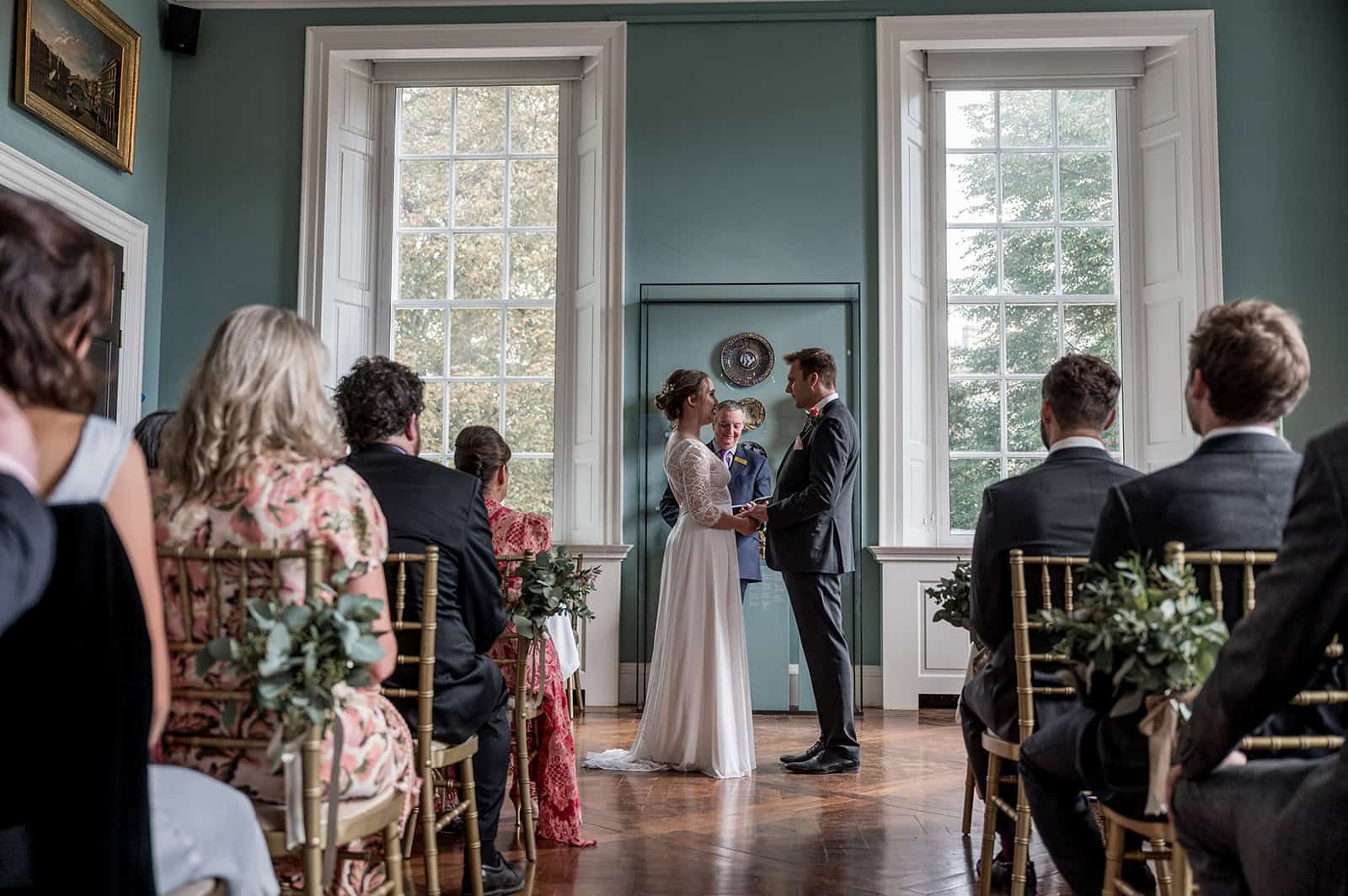 Photo showing a couple getting married in one of the museum galleries, with large windows behind them and gold framed paintings on the walls
