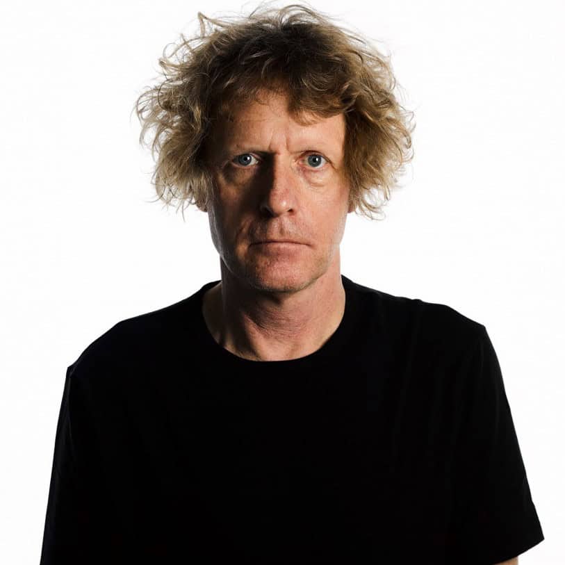 Photograph of artist Grayson Perry wearing a black tshirt with wild hair, against a white background. 