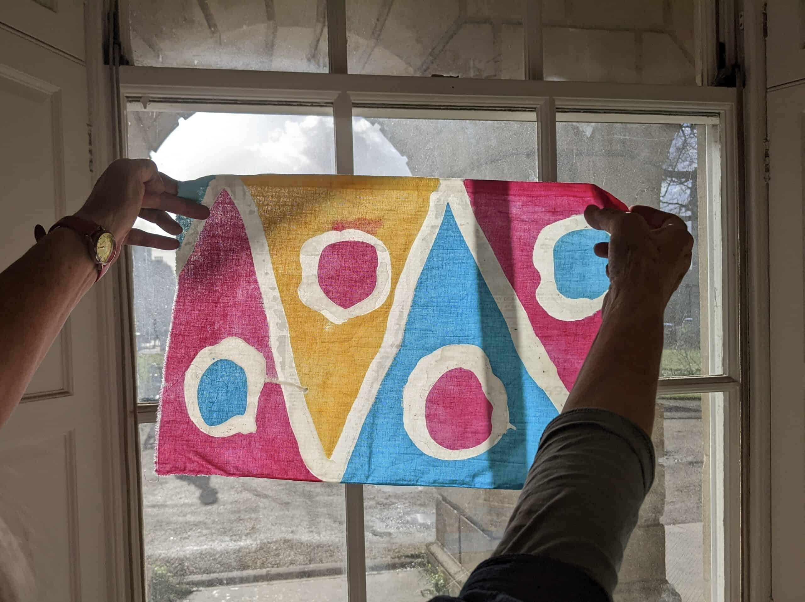 A decorative artwork held up to a window. With a triangular pattern, in the colours pink, yellow and blue.