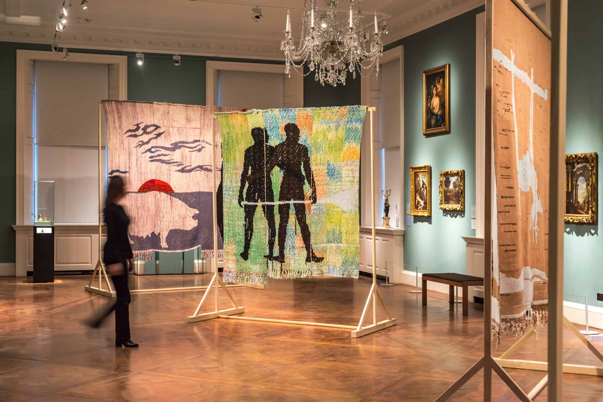 A figure walks through a gallery with large scale colourful weavings by the artist Diedrick Brackens