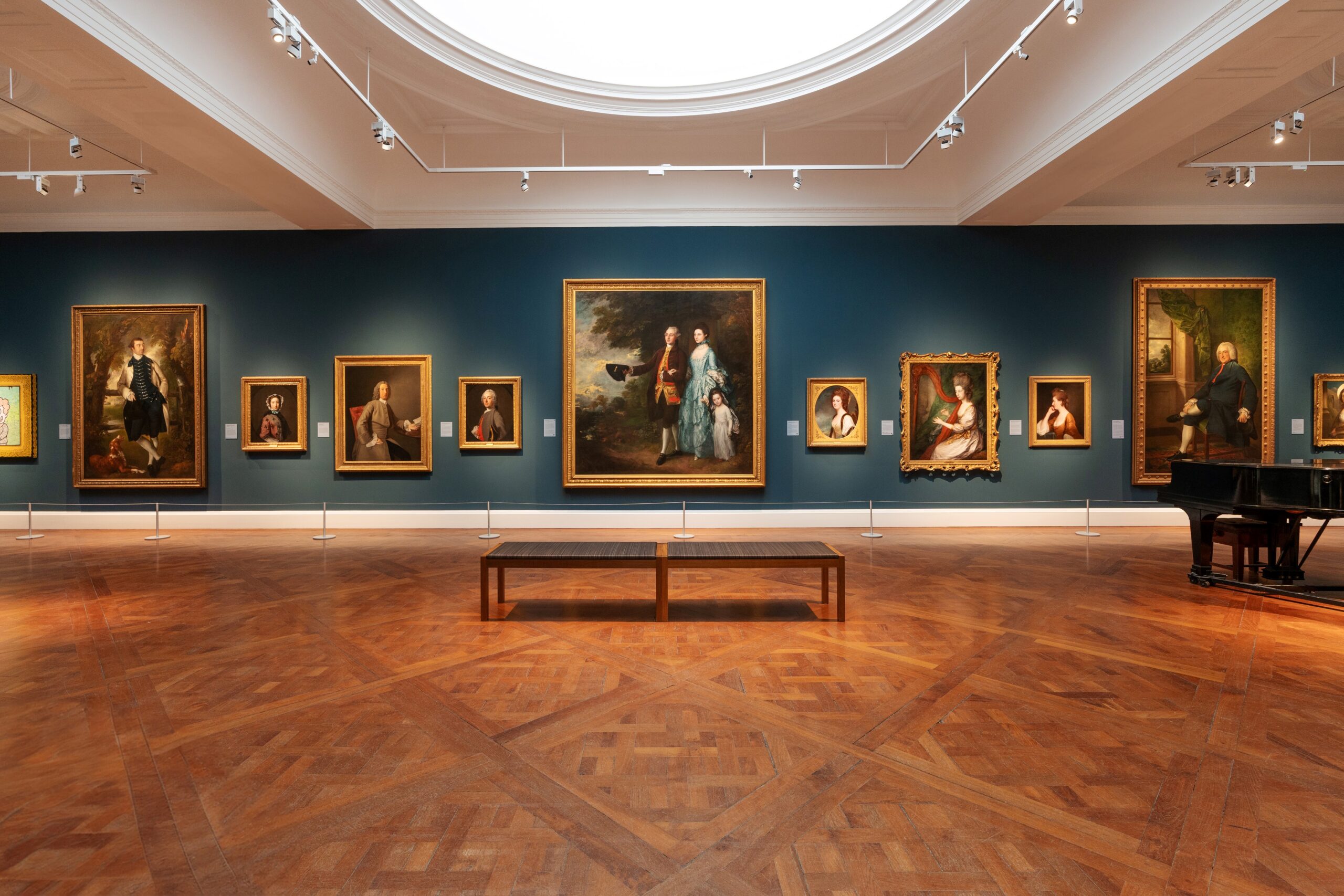 Photo showing a gallery at the Holburne Museum with dark blue walls, wooden floor and portrait paintings in gold frames hung in a row. There is also a grand piano, and a bench for sitting on.
