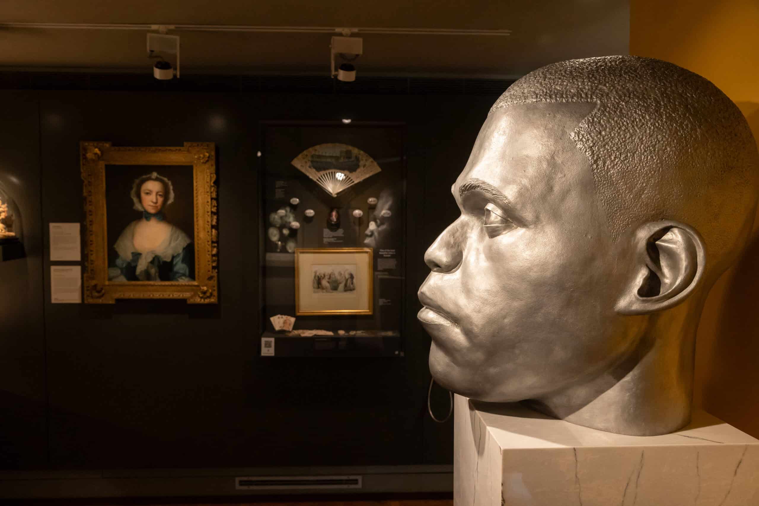 Photo showing a large sculptural head in the foreground, with historic paintings and decorative objects displayed behind
