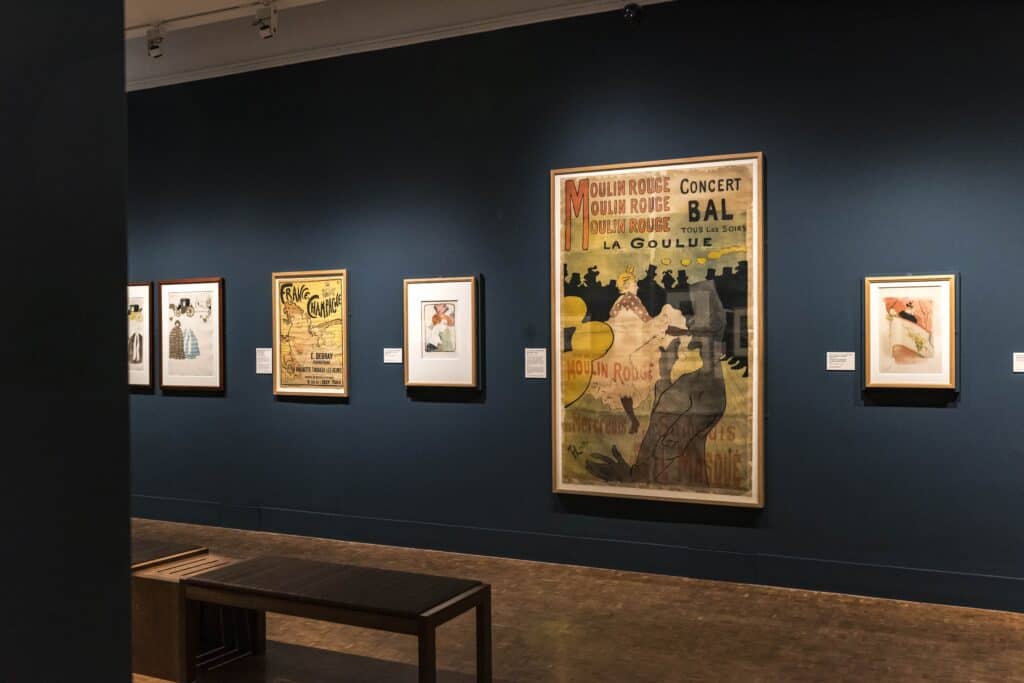 Photo showing a gallery space with dark blue walls, hung with several prints including a large scene of a can-can dancer