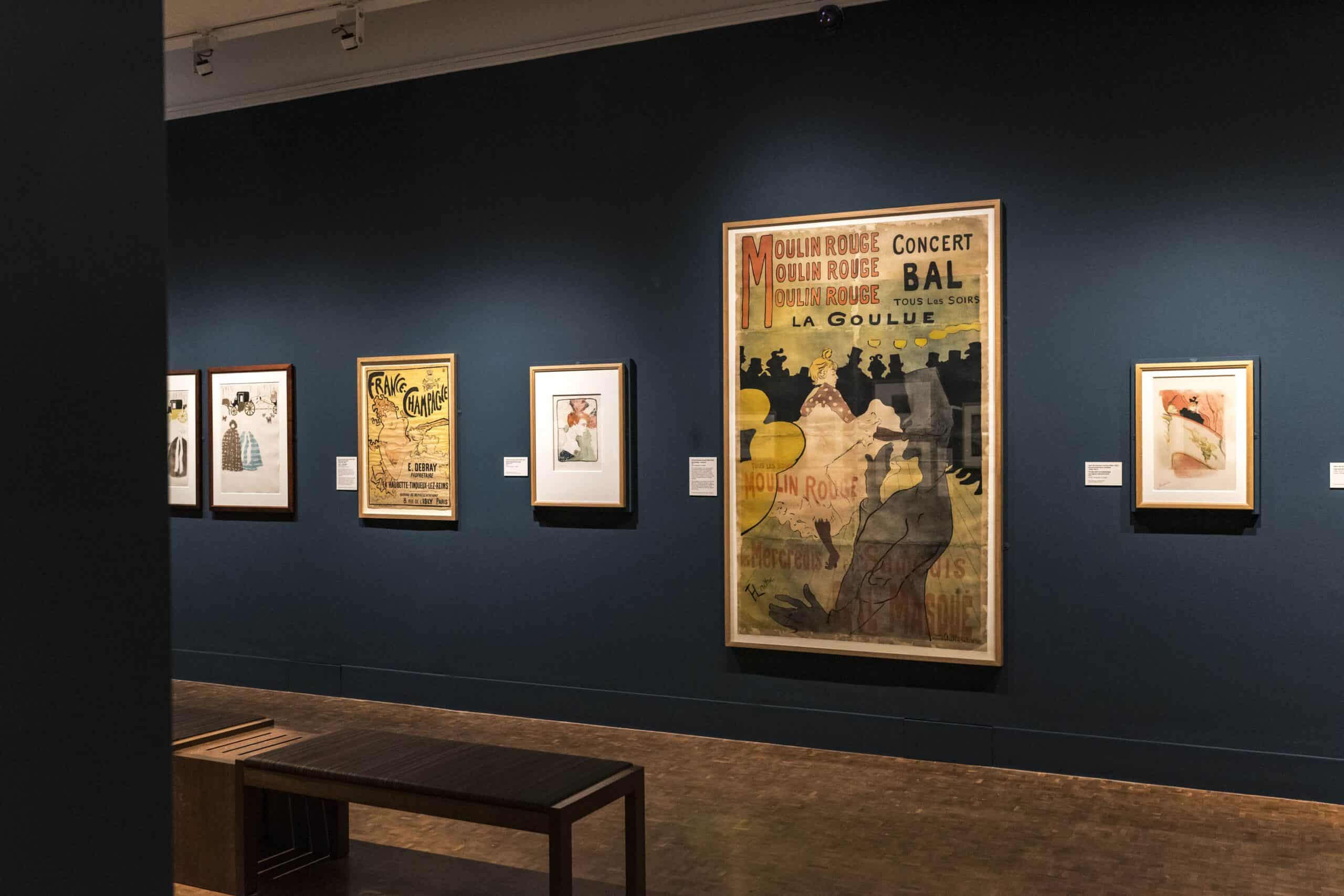 Photo showing a gallery space with dark blue walls, hung with several prints including a large scene of a can-can dancer
