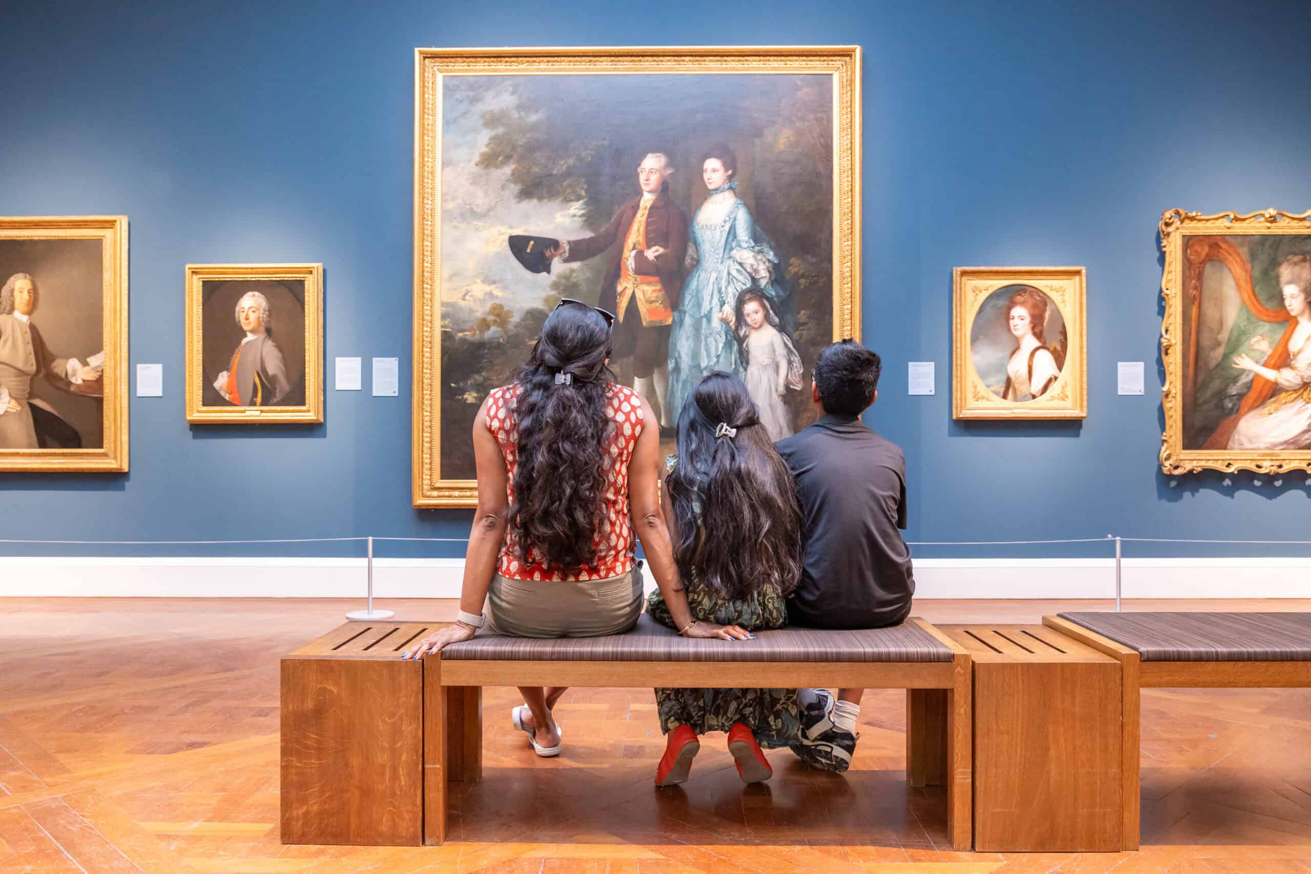 A family sit on a bench looking at large paintings in ornate gold frames against a dark blue wall