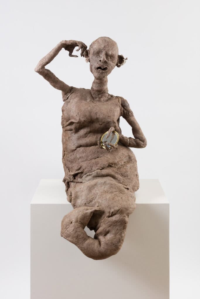 Photo showing a sculptural form of a mermaid figure created from rough textured rubber, clutching a silver mirror in its hand